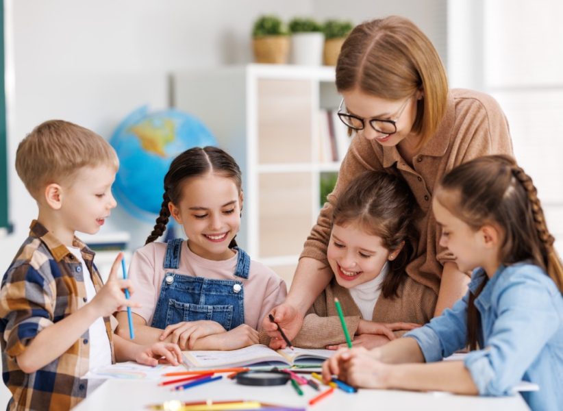 Smart woman teacher in glasses helping students to to schoolwork during lesson in light classroom at school