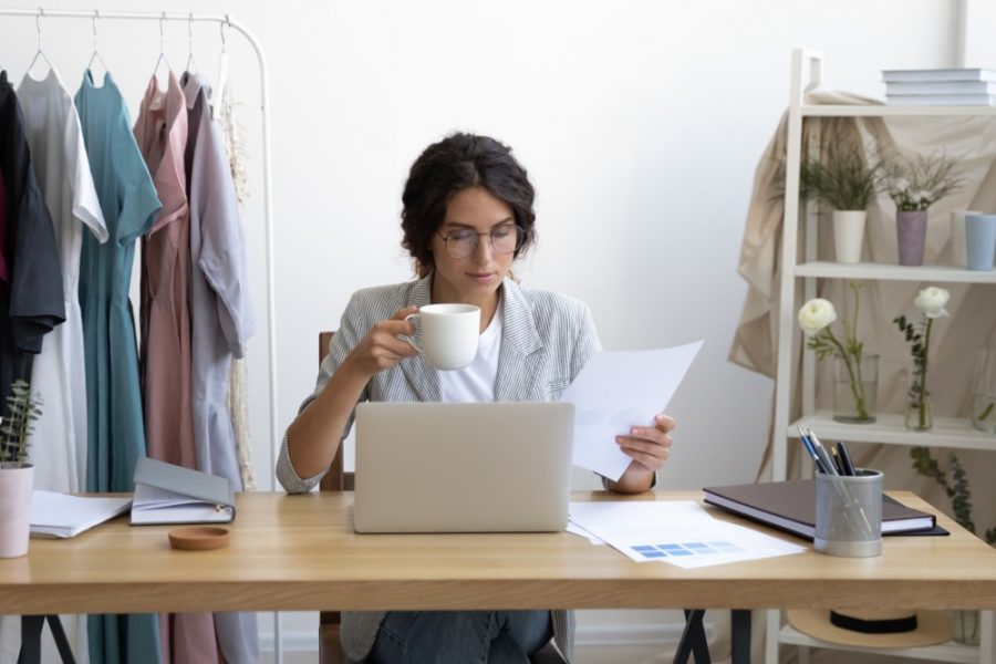 Attractive young businesswoman drinking morning cup of hot coffee, looking through financial paper documentation at workday beginning in office, happy designer enjoying break time at workplace.